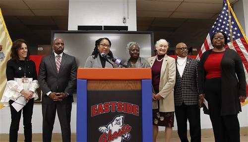 Several women stand behind a podium while the one in the middle makes comments into the microphone.